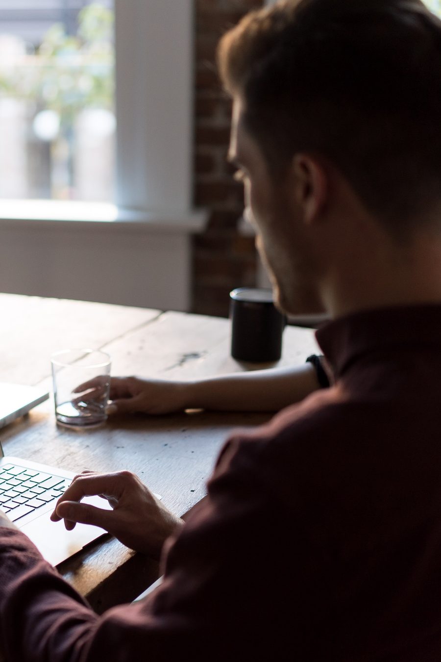 A man in a dark red jumper typing on a laptop at a wooden table by a window, a glass and a mug beside him