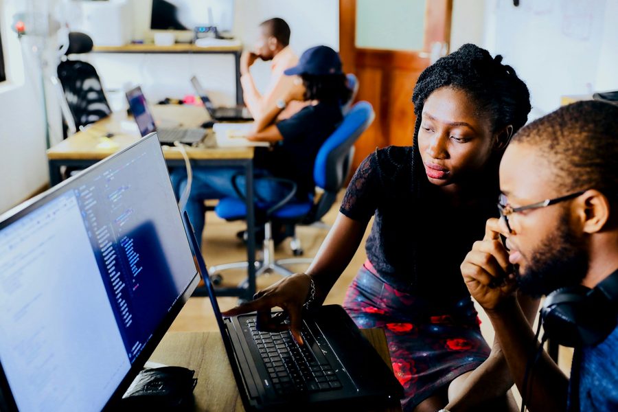 Two colleagues at a desk reading code, one reaching for the laptop keyboard while the other watches the monitor beside it