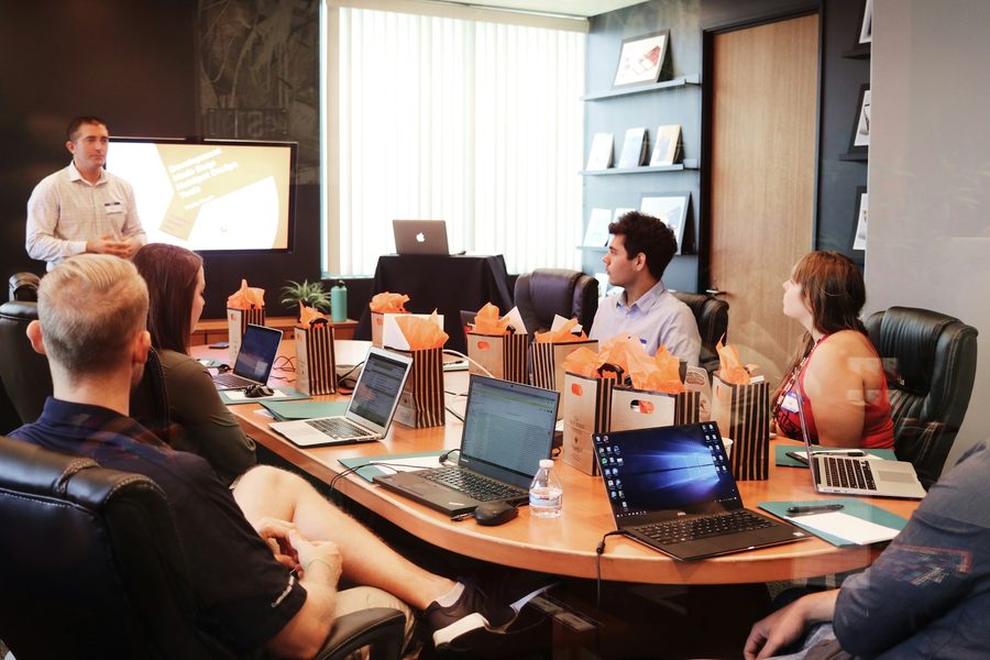 A presenter standing beside a wall mounted screen while colleagues follow from laptops around a boardroom table