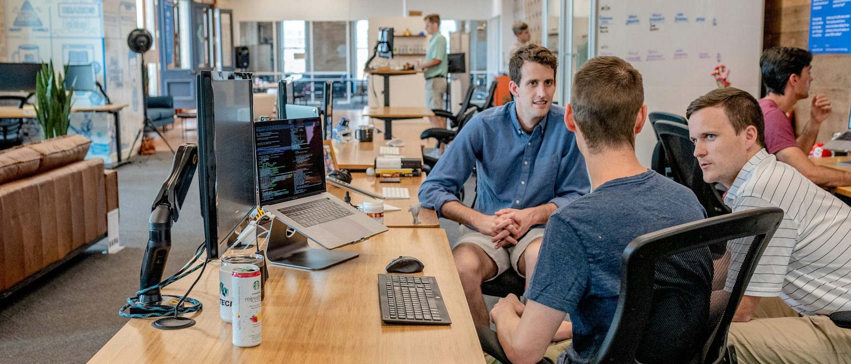 Three colleagues talking beside a desk in a long open plan office, a monitor showing code in the foreground and whiteboards on the wall behind