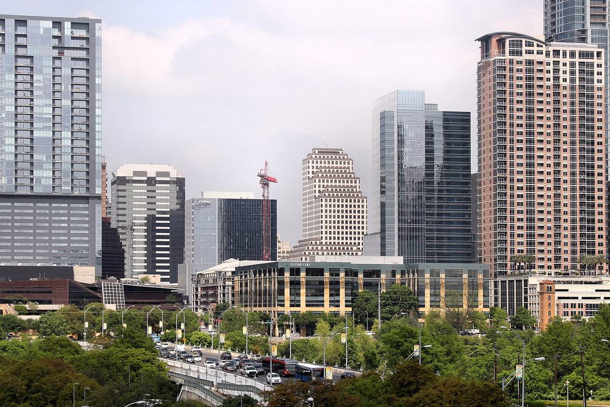Downtown Austin office and apartment towers seen from the south on an overcast day, with traffic crossing an elevated road behind a line of trees