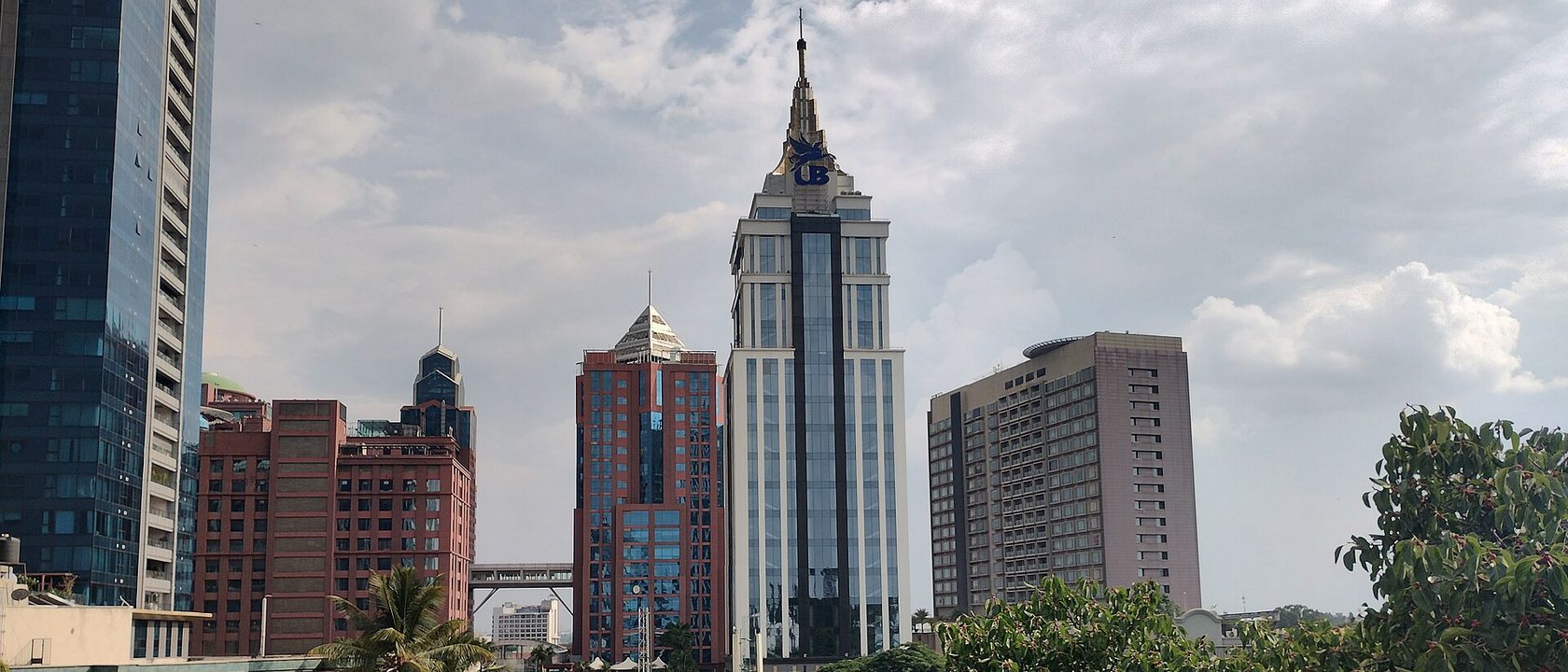 The UB City towers on Vittal Mallya Road in Bengaluru under a cloudy sky, a spired white and glass tower flanked by red brick office blocks and palm trees