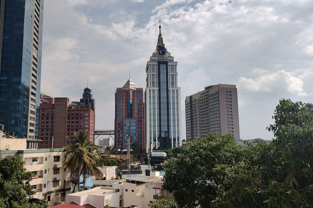 The UB City towers on Vittal Mallya Road in Bengaluru in daylight, a spired white and glass office tower rising above red brick blocks, apartment roofs and trees