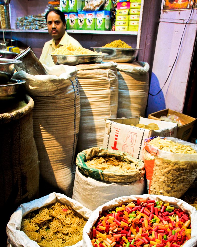 A shopkeeper standing behind open sacks of snacks and dried goods in a small Indian shop, packets stacked on the shelves behind him