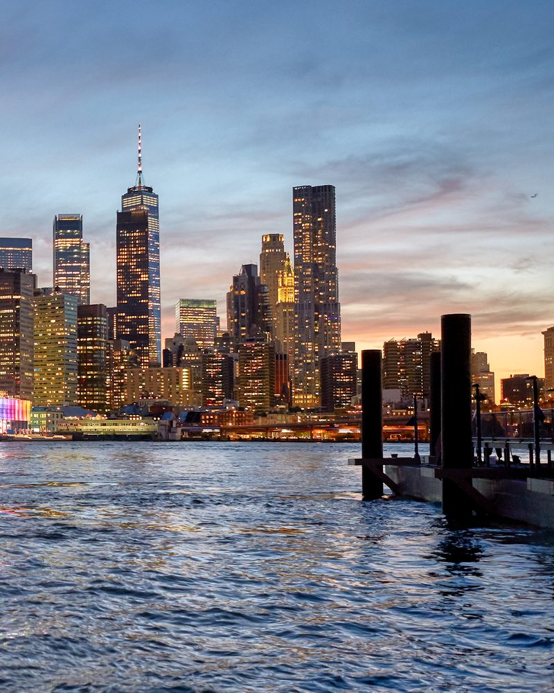 The Lower Manhattan financial district at dusk seen across the East River, One World Trade Center rising above lit office towers with mooring piles in the foreground