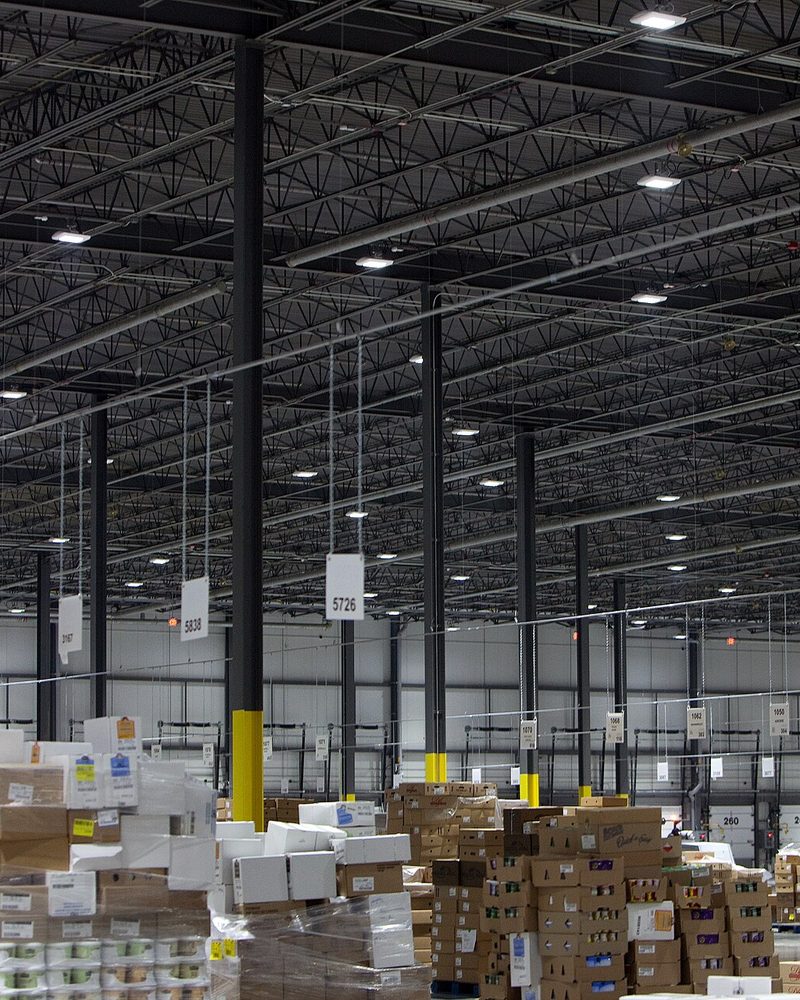 The inside of a distribution warehouse, pallets of stacked cartons on the floor under a wide steel roof hung with numbered bay markers