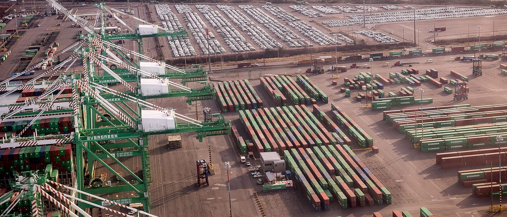 Aerial view of a container terminal, green ship to shore cranes standing over rows of stacked shipping containers with a car storage yard beyond