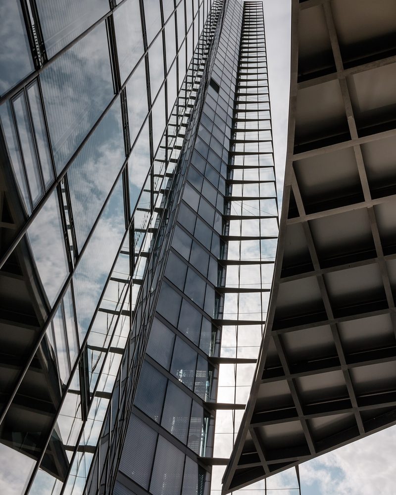 Looking up the mirrored glass facade of an office tower against a clouded sky, a concrete coffered soffit cutting across the frame