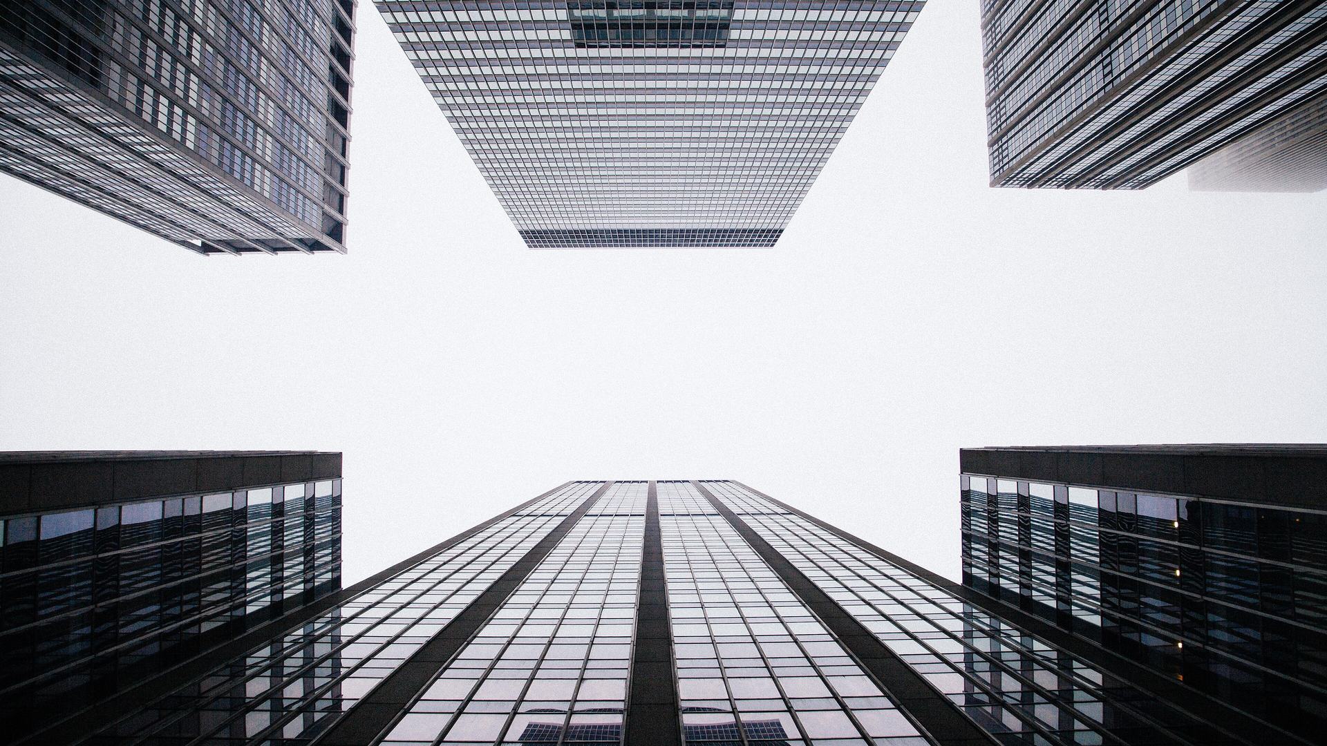 Glass office towers photographed straight up from street level, their facades converging on a pale overcast sky