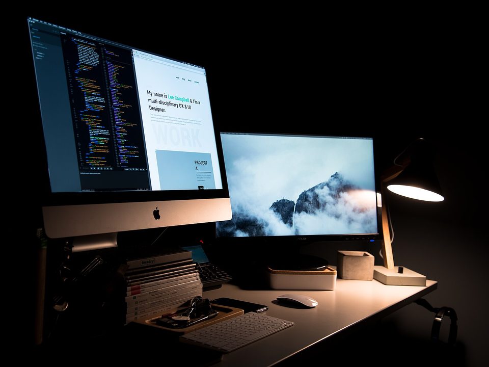 A two monitor desk at night, the left screen split between an open code editor and a web page, lit by a single desk lamp
