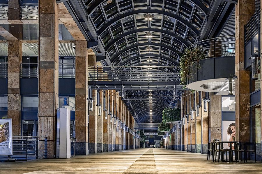 The glazed barrel vaulted arcade of a shopping centre at night, stone piers and lamps running down both sides of the empty walkway