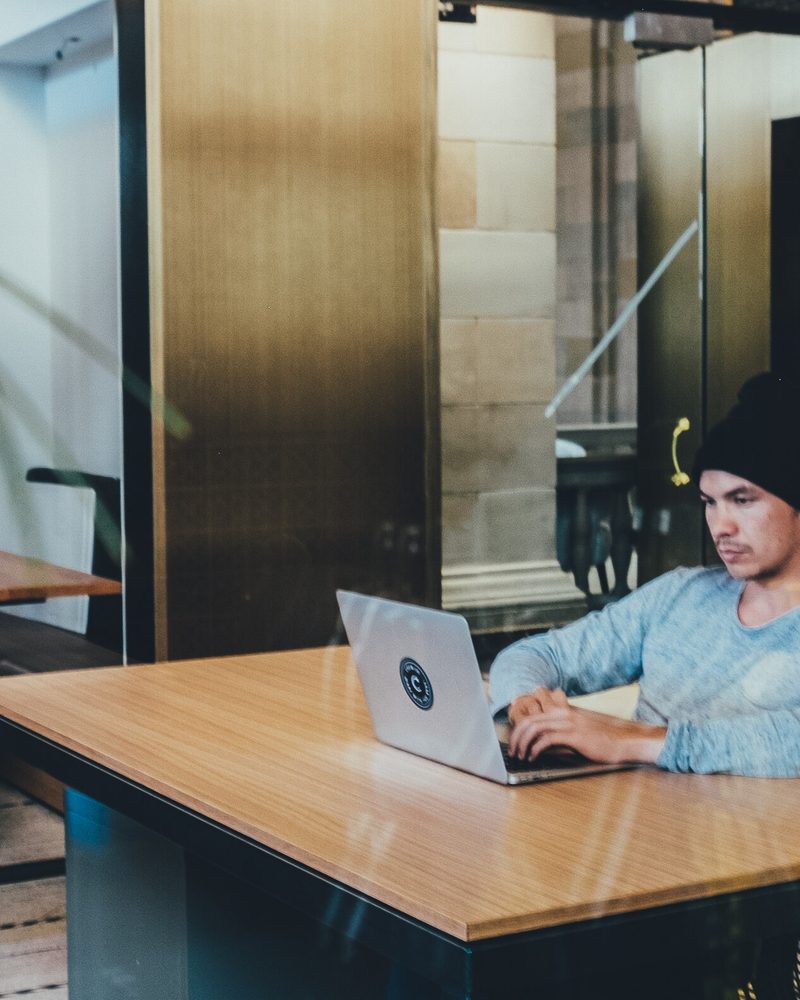 A person in a beanie typing on a laptop at a wide wooden table inside a glass walled workspace